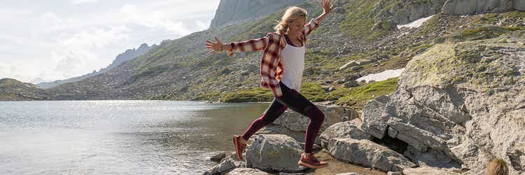 Eine Frau mittleren Alters hüpft lachend von Stein zu Stein, während sie die Arme auf beiden Seiten wegstreckt. Die Steine sind im seichten Wasser eines Sees.