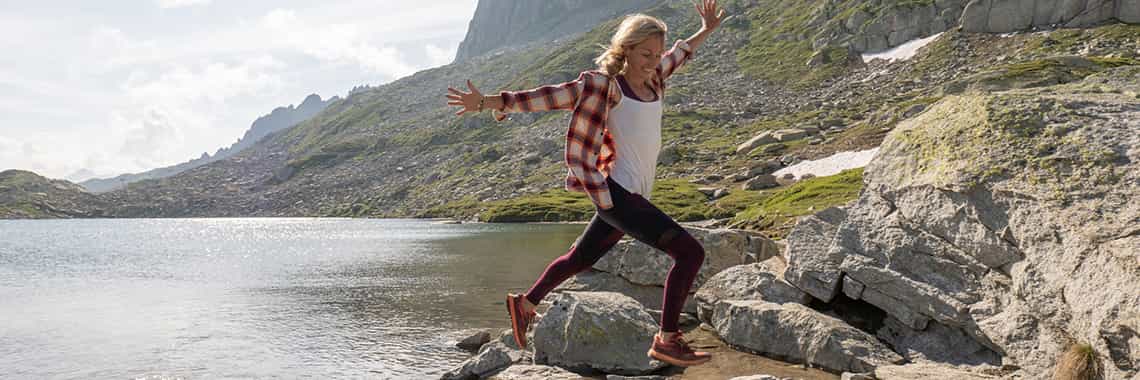 Eine Frau mittleren Alters hüpft lachend von Stein zu Stein, während sie die Arme auf beiden Seiten wegstreckt. Die Steine sind im seichten Wasser eines Sees.
